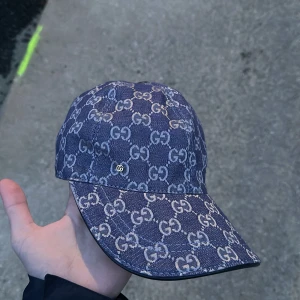 Blå jeanskeps från Gucci med GG-mönster - Säljer en blå keps från Gucci i jeansmaterial med klassiskt GG-mönster över hela kepsen. Justerbar rem baktill i svart läder och vit insida. Perfekt för dig som gillar streetwear och vill sticka ut.