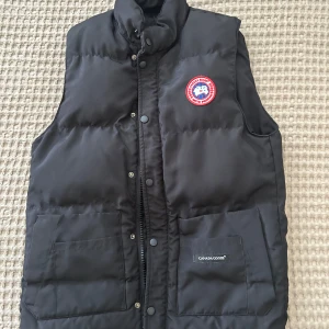 Svart dunväst från Canada Goose - Säljer en svart dunväst från Canada Goose med klassisk logotyp på bröstet. Västen har hög krage, tryckknappar och dragkedja framtill samt två stora fickor. Perfekt för kyliga dagar.