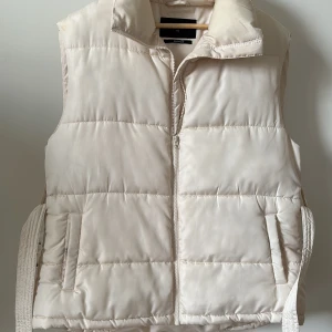 Vit dunväst från Bik Bok - Snygg vit dunväst från Bik Bok i storlek XS. Västen har en dragkedja framtill och är perfekt för kyligare dagar. Den quiltade designen ger en stilren look och den är tillverkad av återvunnet material. Västen har också smink fläckar man ser det på sista bilden om man kollar noga.