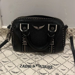 Zadig & Voltaire sunny nano väska - Cool svart handväska från Zadig & Voltaire i skinn med silverfärgade metalldetaljer och nitar längs kanterna. Väskan är köpt här på plick så har ändåst påsen, väskan är lite rostig på få ställen som ni ser på bilderna (därav bra pris) men annars i fint skick! Skriv vid frågor och funderingar!💕