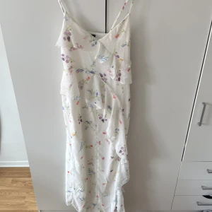 Vit blommig långklänning med volang - Superfin vit långklänning med tunna axelband och blommigt mönster i lila, blått och rosa. Klänningen har volangdetaljer framtill och längs kjolen, vilket ger en romantisk känsla. Perfekt för sommarens alla tillfällen!