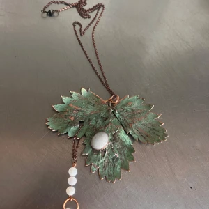 Halsband med grönt blad och pärldetaljer - Unikt halsband med ett stort grönt blad i metall och kopparfärgad kedja. Framsidan har en vit pärla och flera små pärlor hänger i en kedja. Baksidan har en dekorativ koppartråd. Perfekt för dig som gillar naturinspirerade smycken.