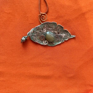 Handgjort halsband med lövformad hänge - Unikt halsband med ett lövformat hänge i silverfärgad metall och en grön sten i mitten. Hänget har fina detaljer och hänger i ett brunt band. Perfekt för dig som gillar naturinspirerade smycken.