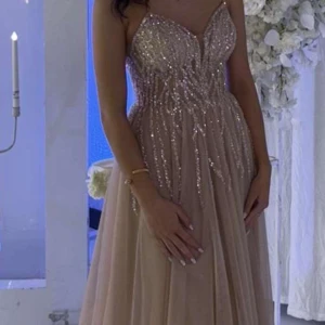 Glittrig champagnefärgad långklänning - Säljer en superfin långklänning i champagnefärg med glittriga paljetter upptill och v-ringad urringning. Klänningen har  ett flowy fall, perfekt för speciella tillfällen. Storlek går att justera med snörning