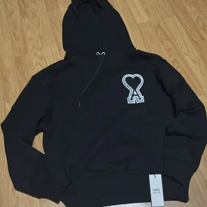 Svart hoodie från Ami med broderad logga - Säljer en svart hoodie från Ami med stor broderad logga i vitt och grått på bröstet. Tröjan har huva med dragsko och ribbade muddar. Perfekt för en avslappnad och stilren look. (Hör av er vid minsta fundering, PRISET ÄR INTE HUGGET I STEN)🤝