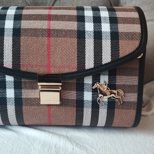 Rutig axelväska från Burberry - Säljer en snygg rutig axelväska från Burberry med klassiskt mönster i beige, svart, vitt och rött. Väskan har guldfärgad kedja, metallspänne och en dekorativ hästdetalj. Sidorna är i trälook med Burberry-logga.