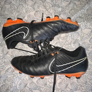 Nike svarta fotbollsskor med orange sula - Säljer ett par svarta Nike fotbollsskor med orange sula och dobbar. Skorna har klassisk snörning och en stor vit Nike-logga på sidan. Ovandelen är i syntetmaterial och designen är sportig och clean, perfekt för dig som vill sticka ut på planen.