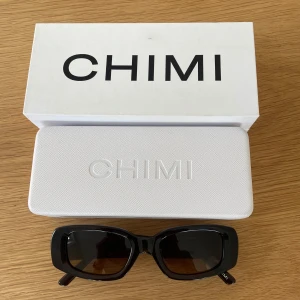 Bruna solglasögon från CHIMI - Snygga bruna solglasögon från CHIMI med rektangulära bågar och mörkbruna glas. Bågarna är i blank plast och modellen är stilren och modern. Perfekta för dig som vill ha en clean och trendig look. Kommer med originaletui och box.