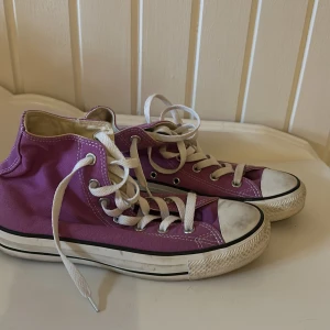 Lila Converse Chuck Taylor All Star - Säljer ett par klassiska Converse Chuck Taylor All Star i lila canvas med vita snören och gummitå. Skorna har högt skaft, rund tå och den ikoniska loggan på sidan. Perfekta för dig som vill ha en färgklick till outfiten. Använda en handfull tillfällen pga för stora för mig.