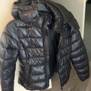 Svart pufferjacka med huva - Säljer en svart pufferjacka med glansig finish och huva. Jackan har dragkedja framtill, två fickor med dragkedja och quiltad design. Perfekt för kalla dagar när du vill hålla stilen och värmen.