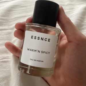 ESSNCE Warm'n Spicy Eau de Parfum - Parfymflaska från ESSNCE med doften Warm'n Spicy. Flaskan är cylindrisk i genomskinligt glas med svart rund kork och vit etikett med svart text. Modern och stilren design, perfekt för dig som gillar varma och kryddiga dofter.