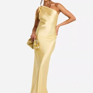 Fin gul långklänning i satin - Supersnygg långklänning i champagnefärgad satin med oneshoulder och knyt-detalj på axeln. Klänningen har en elegant, slät siluett som faller snyggt längs kroppen. Perfekt för dig som vill sticka ut med en lyxig och trendig look.