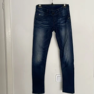 G-Star Lynn Skinny jeans blå 27/32 - Snygga G-Star Lynn Skinny jeans i mörkblå tvätt med slitningar och klassiska fem fickor. Jeansen har skinny fit, låg midja och är tillverkade i stretchig denim för en bekväm passform. Perfekta för dig som gillar en smal siluett och cool streetstil.