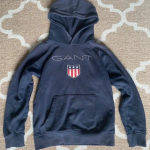 Mörkblå hoodie från GANT med logga - Mörkblå GANT hoodie i storlek 134/140. Ganska gammal så lite sliten (skriv för mer bilder på de) men inget man lägger så mycket märke till. På lappen inne i tröjan där det står hur man tvättar står även namnet på tidigare ägaren men inte så tydligt och inget man ser när man har på sig tröjan (skriv för bilder på de) Nypris ca 550kr! Pris går att diskutera 