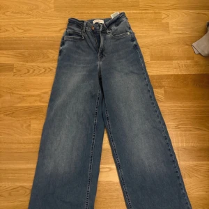 Blå wide jeans med hög midja - Säljer ett par blå jeans med hög midja och riktigt breda ben. Klassisk femficksmodell med knapp och dragkedja framtill. Jeansen har en snygg tvätt och raka, vida ben för en trendig look. Perfekta till sneakers eller boots.