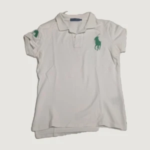 Vit piké från Ralph Lauren - Vit pikétröja från Ralph Lauren med klassisk krage och korta ärmar. Stort grönt broderat logomärke på bröstet och siffra på ärmen. Perfekt för en sportig och stilren look.