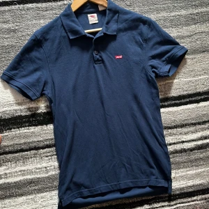  pikétröja från Levi's  - Snygg marinblå pikétröja från Levi's i storlek S. Klassisk krage, kort ärm och två knappar framtill. Liten röd Levi's-logga på bröstet. Perfekt till jeans eller shorts för en stilig och avslappnad look.