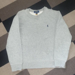 Grå sweatshirt från Polo Ralph Lauren - Klassisk grå sweatshirt från Polo Ralph Lauren med rund halsringning och broderad marinblå logga på bröstet. Tröjan har ribbade muddar vid ärmslut och nederkant, perfekt för en chill och stilren look.