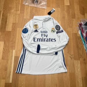 Real Madrid Ronaldo långärmad tröja - Vit långärmad Real Madrid-tröja från Adidas med mörkblå detaljer, FIFA World Champions 2018-märke, Champions League-patchar och Ronaldo 7 på ryggen. Climalite-material som andas och klassisk krage. Perfekt för dig som älskar fotboll och Ronaldo.