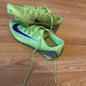 Nike Mercurial fotbollsskor neongula - Säljer ett par Nike Mercurial fotbollsskor i en riktigt fet neongul färg med svarta och blå detaljer. Skorna har snörning och är designade för att ge grymt grepp på planen. Perfekta för dig som vill sticka ut och ha max kontroll på bollen.