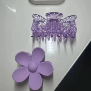 Två lila hårklämmor  - Säljer två superfina lila hårklämmor! En är formad som en blomma i matt plast och den andra har en glittrig, kristall-liknande design med geometriskt mönster. Perfekta för att piffa upp frisyren och ge en söt touch till din look.