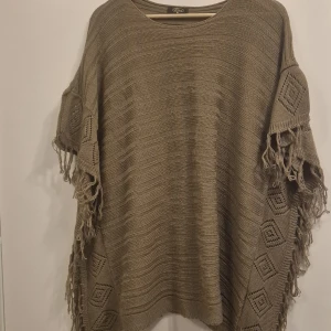 Beige stickad ponchotröja från Elitzia - Säljer en oversized beige stickad ponchotröja från Elitzia. Tröjan har snygga virkade detaljer med diamantmönster på ärmarna och fransar längs kanterna. Perfekt för lager-på-lager och chill höst- eller vårstil.
