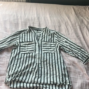 Randig skjorta från Vero Moda M - Snygg vit och svart randig skjorta från Vero Moda i storlek M. Skjortan har v-ringning, två stora bröstfickor och mellan långa ärmar. Perfekt för dig som gillar en avslappnad men trendig look. Materialet känns lätt och luftigt. 