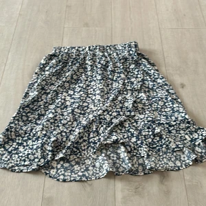 Blommig volangkjol i blått och vitt - Supersöt kort kjol med blå botten och vita blommor. Kjolen har volangkant nertill och resår i midjan för en skön passform. Perfekt för dig som gillar en feminin och lekfull stil.