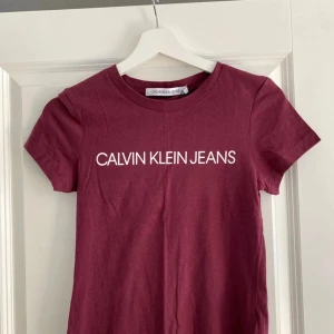 Vinröd t-shirt från Calvin Klein Jeans - Snygg vinröd t-shirt från Calvin Klein Jeans med vit logga tryckt framtill. Klassisk rund halsringning och korta ärmar. Perfekt basic-plagg i mjuk bomull som passar till allt. Enkel och stilren design med normal passform.
