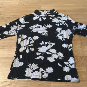 Svart blommig polotopp Gina Tricot - Svart polotopp från Gina Tricot med vita blommor över hela toppen. Toppens material är stretchigt och lätt, med lång ärm och smal passform. Perfekt för dig som gillar en stilren men ändå trendig look.