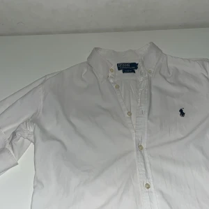 Vit skjorta från Polo Ralph Lauren - Klassisk vit skjorta från Polo Ralph Lauren med broderad marinblå logga på bröstet. Skjortan har lång ärm, knappar framtill och button down-krage. Snygg passform i slim fit och tillverkad i mjuk bomull. Perfekt för en clean och stilren look. Använd en gång, som ny!