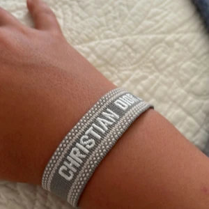 Grått armband från Christian Dior - Snyggt vävt armband från Christian Dior i grått och vitt med tydlig logga. Armbandet har justerbar knytning med två stora tofsar i ändarna för en cool och trendig look. Perfekt accessoar för att lyfta din outfit.