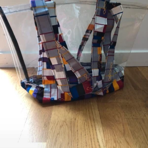 Transparent shoppingväska med färgglada rutor - Stor och rymlig transparent shoppingväska eller beachbag med coola handtag och botten i rutigt mönster med färger som blå, gul, röd och lila. Bandet är tillverkat av unikt mönster i västafrikanskt tyg och den har mjuk plast som skydd för fuktiga handdukar.  Tillverkad i Senegal, oanvänd. Mått: längd 30cm, bredd 15cm och höjd 28cm. 
