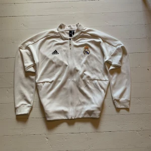 Vit Adidas Real Madrid fotbolls tröja - Fet Real Madrid fotbollströja från adidas. Den är rätt tunn så passar bra som träningströja eller bara att ha till vardags. Den är storlek L. Ända defekten är att den har lite missfärgningar/fläckar på baksidan (se bild 2) men de syns knappt varken på bilderna eller i verkligheten.