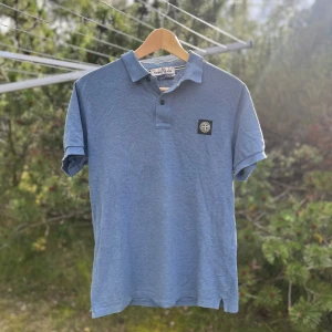 Blå Stone Island pikétröja - Blå piké från Stone Island med klassisk krage, korta ärmar och svart logotyp-patch på bröstet. Tröjan har två knappar vid halsen och är gjord i mjuk bomull. Perfekt för dig som gillar streetwear och vill ha en clean look.
