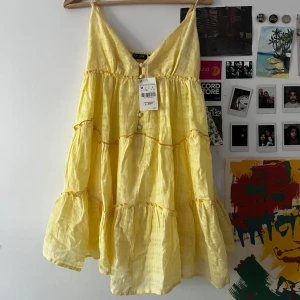 Gullig Gul sommarklänning  - Superfin gul kortklänning från Top n Shop med tunna axelband, v-ringning och knappar framtill. Klänningen har volangiga lager och är gjord i ett lätt, luftigt bomullstyg – perfekt för varma dagar. Söt och flowy stil som ger riktiga sommarvibbar!