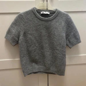 Grå stickad kortärmad tröja från Zara - Mysig grå stickad tröja från Zara med korta ärmar och rund halsringning. Tröjan har en enkel och stilren design med ribbade kanter vid ärmslut och nederkant. Perfekt att styla med jeans eller kjol för en chill look. Köpt förra året använd cirka 5 gånger !🩶🩶 skicka gärna prisförslag 💞