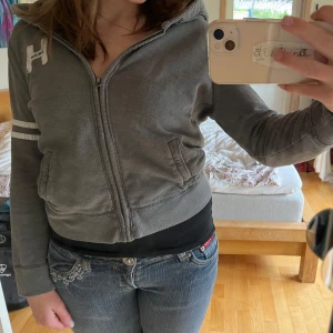 Grå Hollister Hoodie - Säljer en super fin Holliester hoodie i storlek M, passar alla stilar. Den är lite kortare än en ”vanlig” hoodie men det är modellen som ska vara så, den har inga snören vid luvan och ett pyyyytte litet hål vid dragkedjan längst upp (se bild 5) men det märks inte alls, den har inte häller används💗skriv om ni har frågorrr