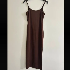 Brun långklänning med slits - En stilren brun långklänning med smala axelband och slits på sidan. Klänningen har en rak passform och ärmlös design, perfekt för en enkel men snygg look. Materialet är mjukt och följsamt, vilket gör den bekväm att bära hela dagen.