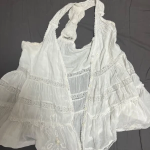 Vit festtopp - Superfin vit linnetopp med broderade detaljer och spetsinslag. Toppen har knappar framtill, volangiga lager och en flätad halterneck. Perfekt för sommarens varma dagar och ger en romantisk boho-vibe. Vintage i superbra skick