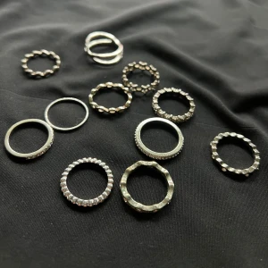 12st silvriga ringar  - Säljer ett set (12st) med flera silvriga ringar i olika former och mönster. Vissa ringar har glittriga stenar, andra har vågiga eller kantiga detaljer och några har stjärnformade utskärningar. Perfekt för att mixa och matcha på olika fingrar. Ej äkta! Storlek S.