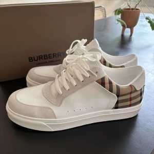 Burberry vita sneakers med rutig detalj - Snygga vita sneakers från Burberry med klassisk rutig panel i beige, svart och rött på hälen. Skorna har ovandel i skinn och mocka, vita snören och en chunky vit sula. Perfekta för dig som vill ha en stilren men ändå ikonisk look.