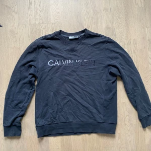 Svart sweatshirt från Calvin Klein - Svart sweatshirt från Calvin Klein med broderad logga på bröstet. Klassisk rund halsringning och långa ärmar. Mjuk bomullskänsla och ribbade muddar vid ärmslut och nederkant. Perfekt för en clean och stilren look.