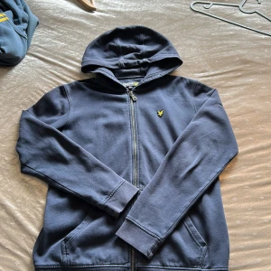 Mörkblå hoodie från Lyle & Scott - Mörkblå hoodie från Lyle & Scott med dragkedja och huva. Klassisk gul logga broderad på bröstet. Tröjan har långa ärmar och två fickor framtill. Perfekt för dig som gillar stilren och enkel design.