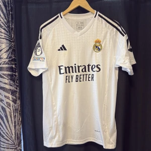 Real Madrid Valverde 8 matchtröja Adidas - Real Madrid fotbollströja med Valverde 8 på ryggen. Vit med svarta detaljer och broderat klubbmärke. Adidas-logga på bröstet, Champions League-märke och Foundation-patch på ärmen. Slim fit-modell i lätt och ventilerande material.