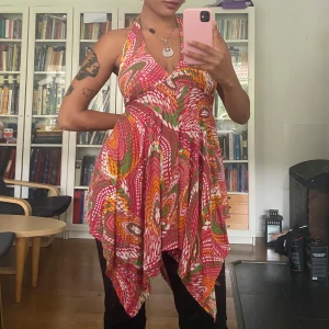 Färgglad halterneckklänning  - Säljer en superfin kortklänning i boho-stil med färgstarkt mönster i rosa, orange, vitt och grönt. Klänningen har halterneck med knyt i nacken, v-ringning och öppen rygg. Materialet känns lätt och flowy, perfekt för varma dagar. Asymmetrisk nederkant för extra edge💥