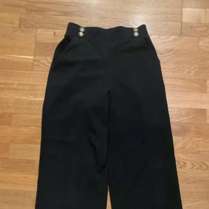 Svarta vida kostymbyxor från River Island - Svarta byxor från River Island med hög midja och vida ben. Snygga detaljer med dubbla knappar i midjan och diskreta fickor framtill. Resår i midjan, vilket gör att den passar flertal storlekar. Perfekta för dig som gillar en stilren och trendig look.