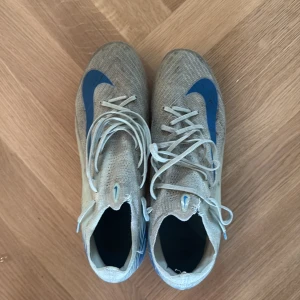 Nike fotbollsskor med blå detaljer - Säljer ett par Nike fotbollsskor i ljusblåa med blå swoosh och grafiska detaljer. Skorna har snörning, platt sula med dobbar och är tillverkade i syntetmaterial. Perfekta för dig som vill ha stilrena och snabba skor på planen. Ag dobbar 