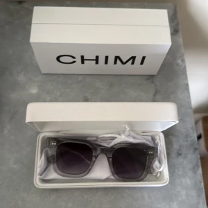 Grå transparenta solglasögon CHIMI - Säljer ett par stilrena solglasögon från CHIMI med transparent grå båge och mörka glas. Rektangulär form som ger en modern vibe. Kommer med originaletui och putsduk. Perfekta för att lyfta din look i sommar! 