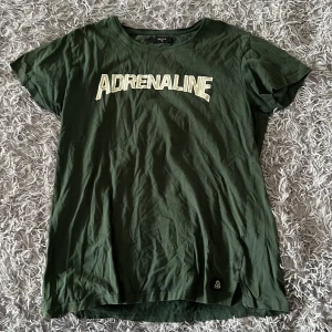 Grön t-shirt från Lager 157 - Mörkgrön t-shirt från Lagar 157 i bomull med coolt ADRENALINE-tryck i vitt/grått på bröstet. Klassisk rund hals och korta ärmar, perfekt för en avslappnad streetstil. Liten etikett nertill. Skön och enkel att matcha med jeans eller shorts. Den har ett pyttelitet hål på ryggen (se bild)